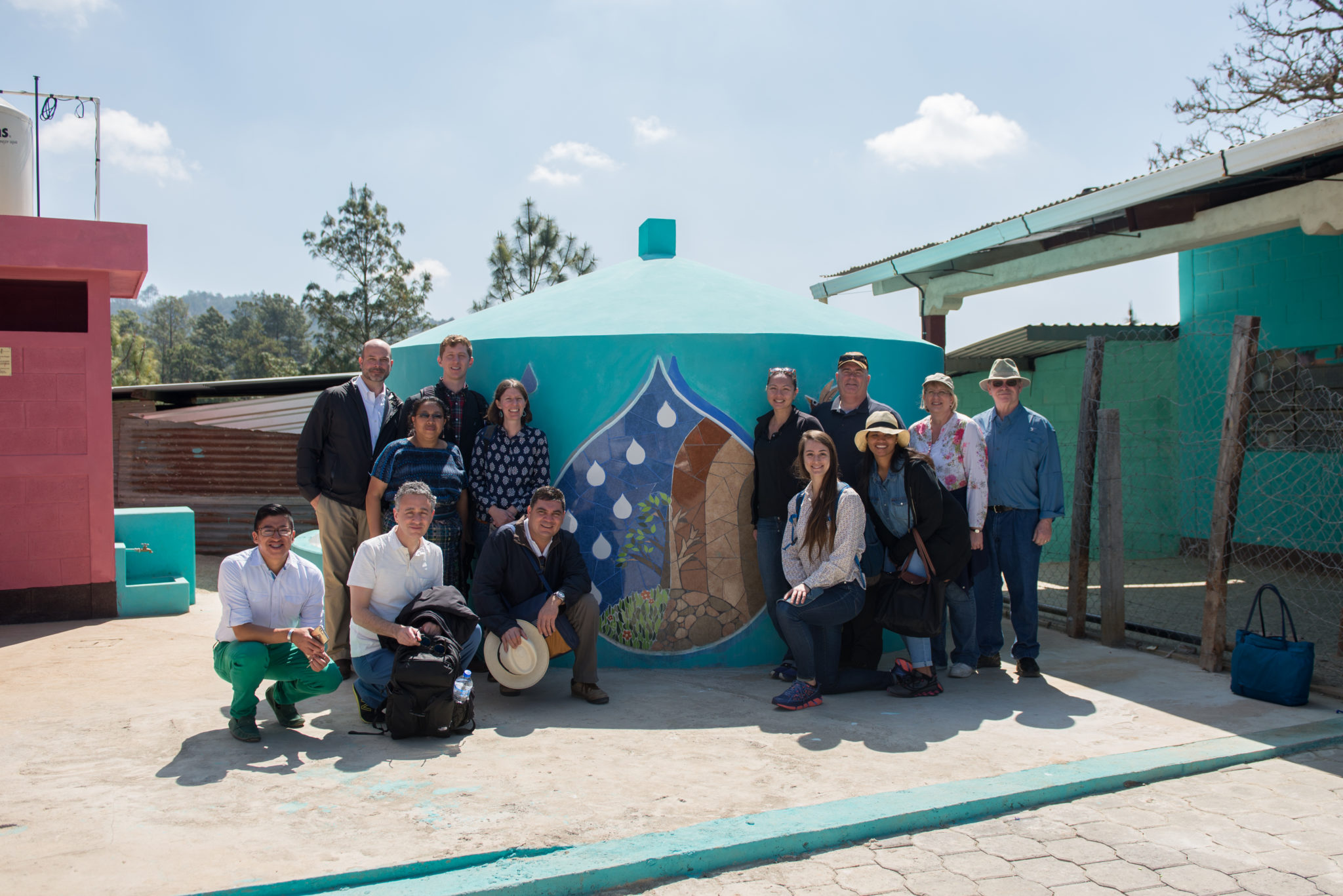Guatemala-SantaCruzdelQuiche-201703-75 Photograph of a group of people standing before a blue watertank with a colorful mosaic