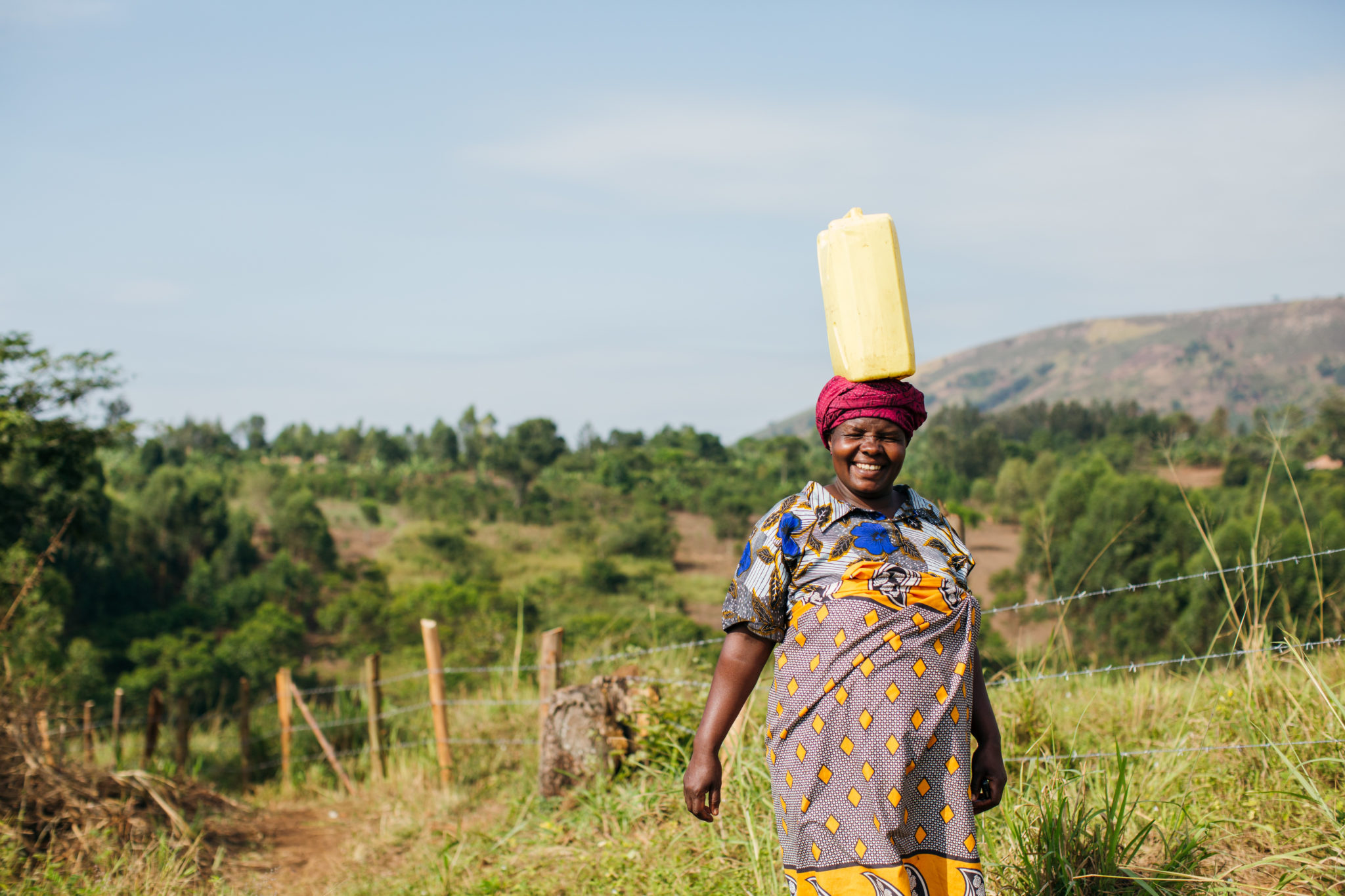 Uganda_2016 (310 de 369) Photograph of a woman in colorful clothing with a water jug balanced on her head.