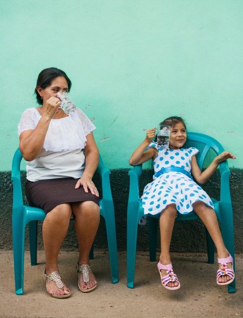 Photo of a mother and daughter seated in front of a teal wall drinking water