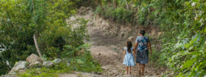 Photograph of a woman and a small child walking away from the camera on a road surrounded by green plants