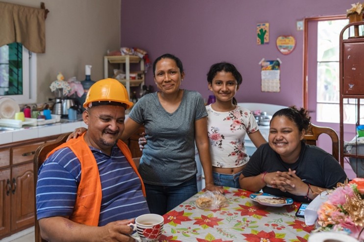 Photograph of a man wearing a safety helmet and vest sitting at a table with his smiling family