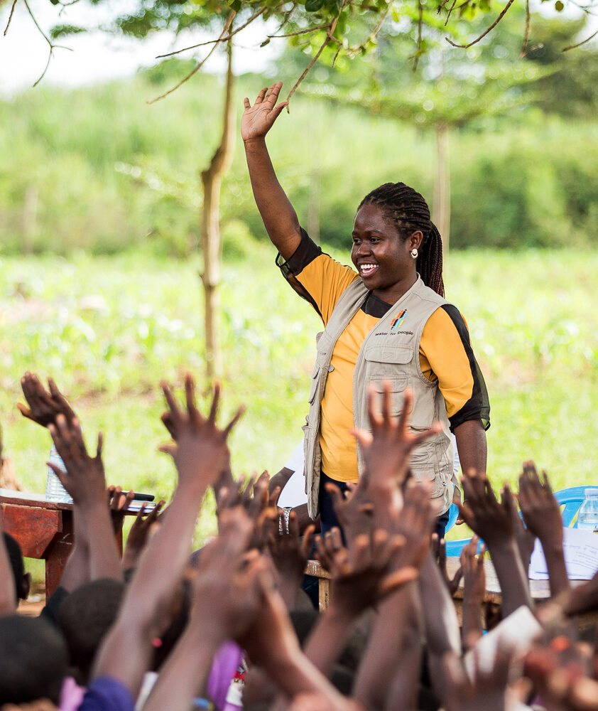 A woman wearing a Water For People vest raises her hand in front of a group of students also raising their hands against a green, outdoor background.