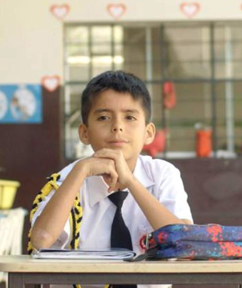 A young boy in a white school uniform with a black tie sits at a desk in a classroom, resting his chin on his hands with a thoughtful expression.