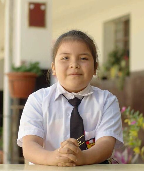 A young girl in a white school uniform with a black tie wits at a desk and smiles for a portrait photo, she is looking straight to the camera.