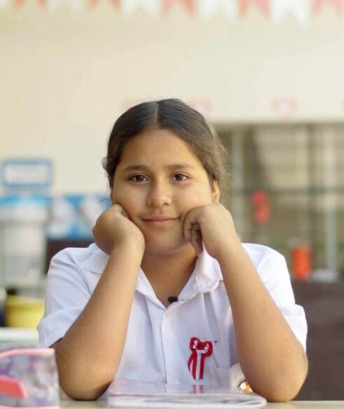 A young girl in a white school uniform sits at a desk, resting her chin on her hands with a gentle smile.