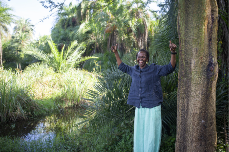 Grace, senior program officer at Water For People Uganda stands next to one of restored wetlands smiling, and with both hands raised up in a celebratory motion.