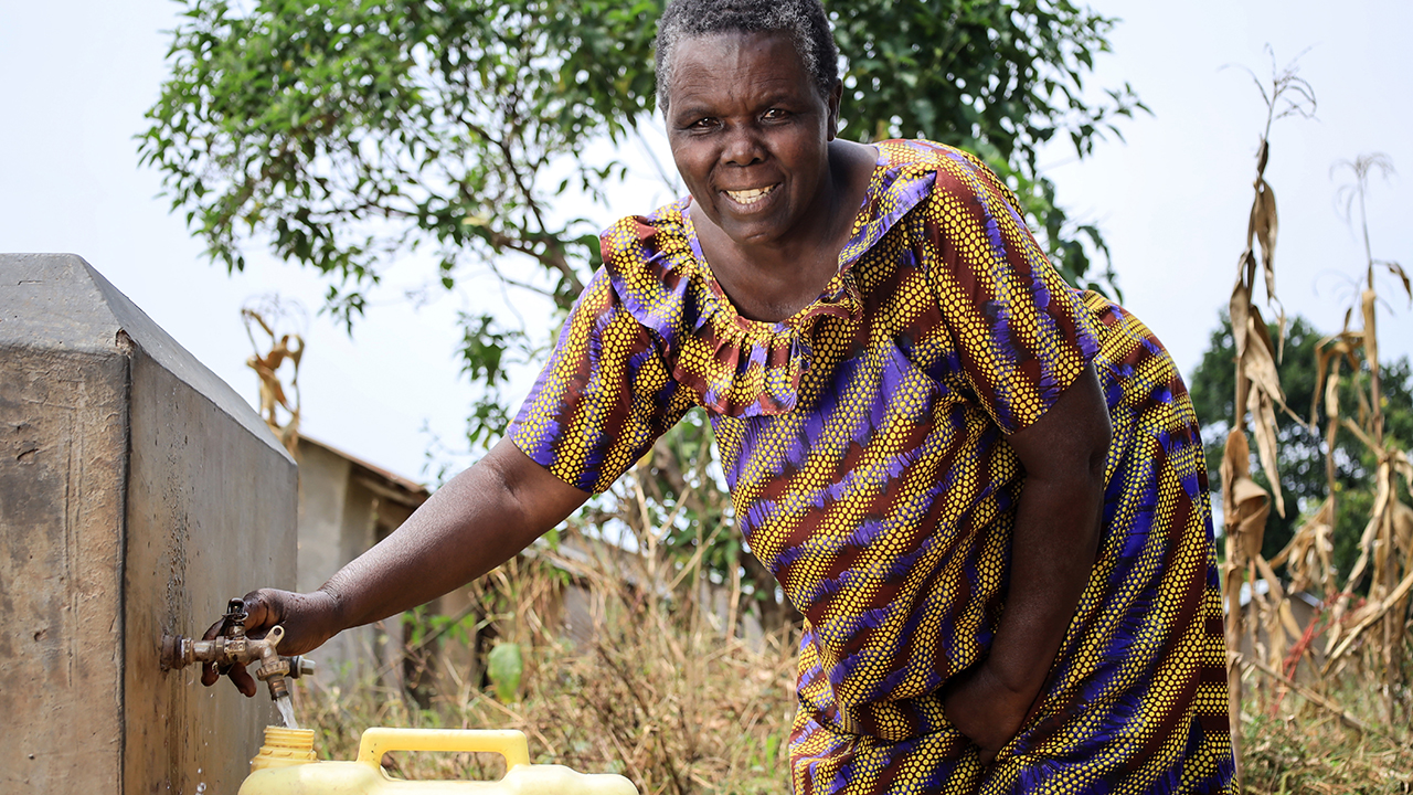 Beatrica, in a vibrant yellow shirt, fills a jerry can of water at a new tap while smiling at the camera.