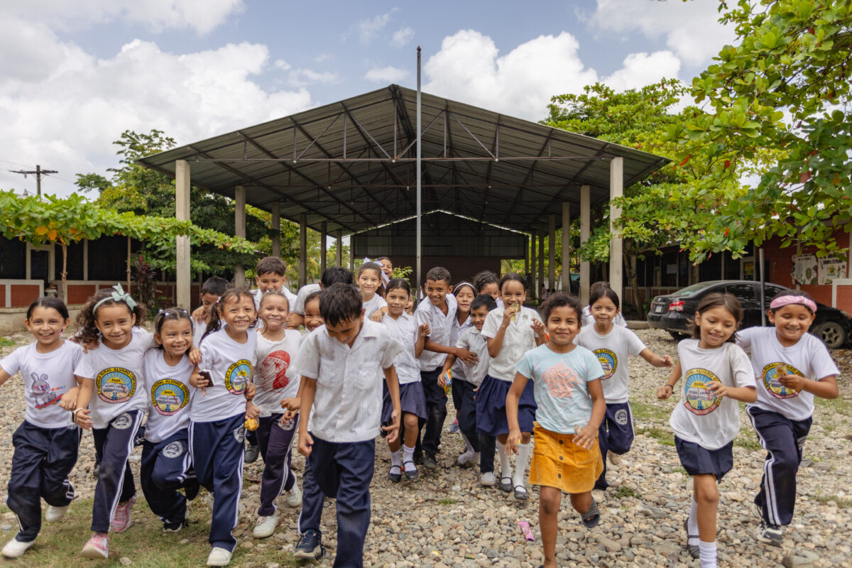 Group of schoolchildren in uniforms running and smiling on a gravel path toward the camera, with a large open-sided school structure and greenery in the background.