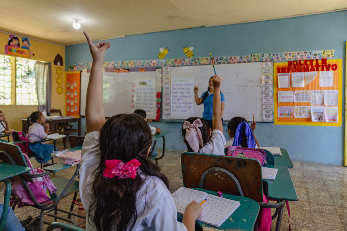 One student raises her hand during class while the teacher points at a poster on a wall about hygiene practices.