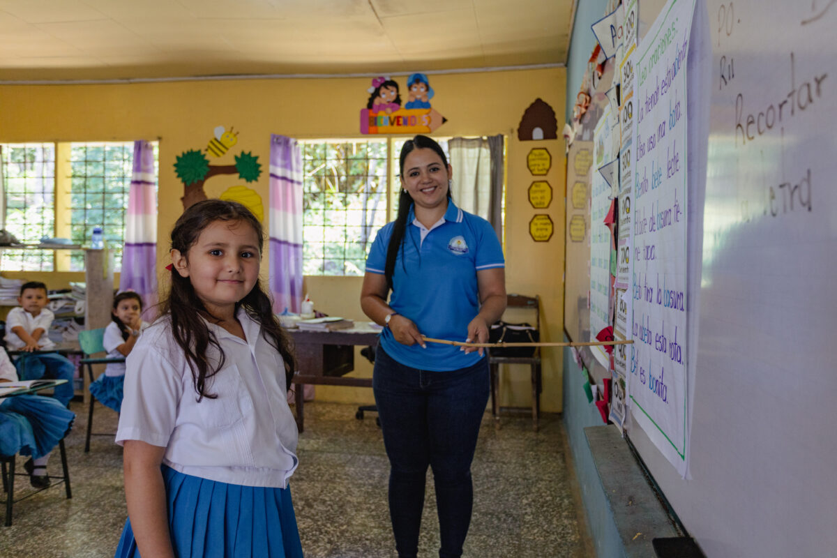 Karla, a teacher at a school in Honduras stands with one of her students while pointing to an illustrated hygiene poster on a wall. More students sit at desks in the background.