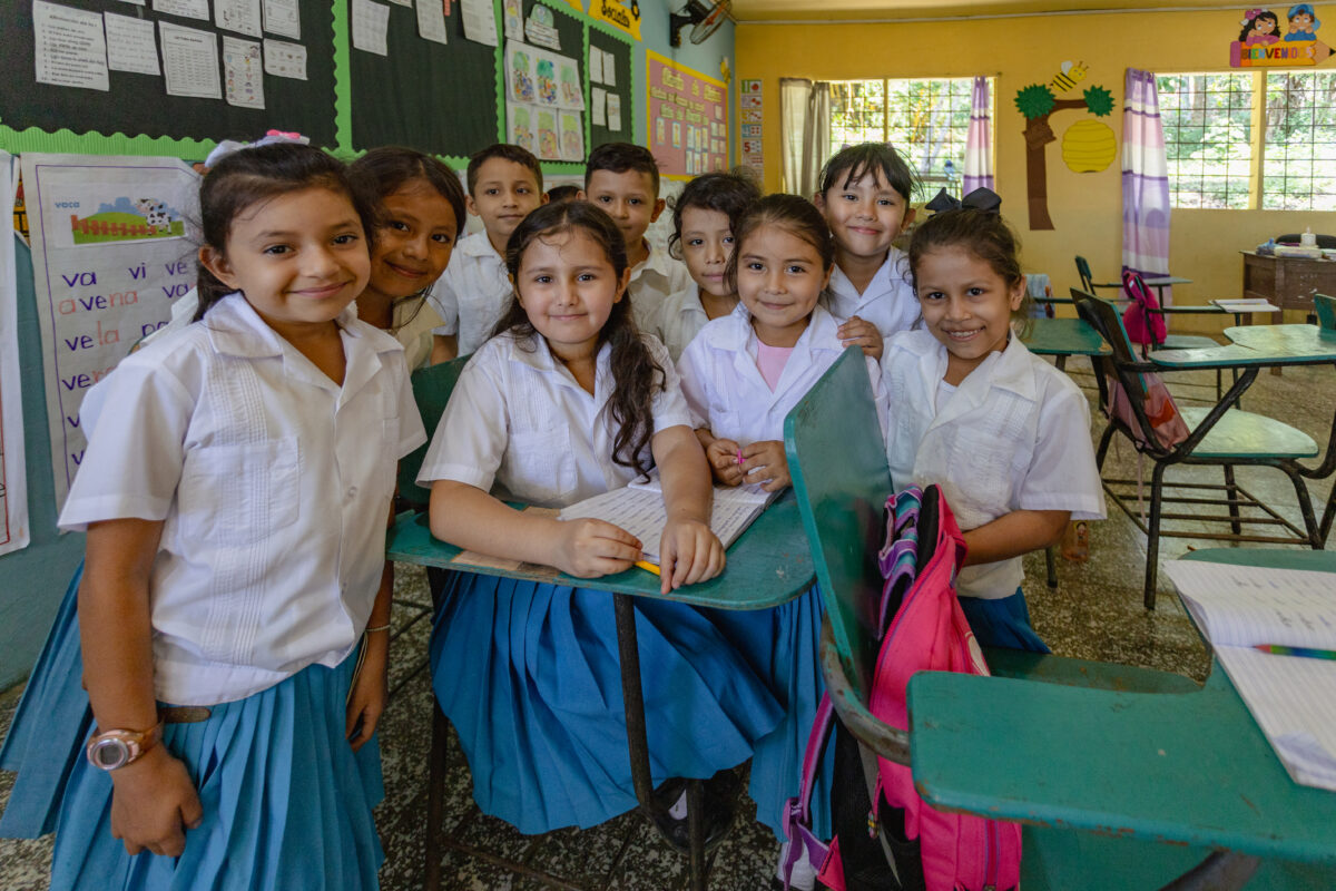 Group of elementary school students in uniforms gathered around desks in a classroom, facing the camera, with colorful posters and learning materials on the walls.