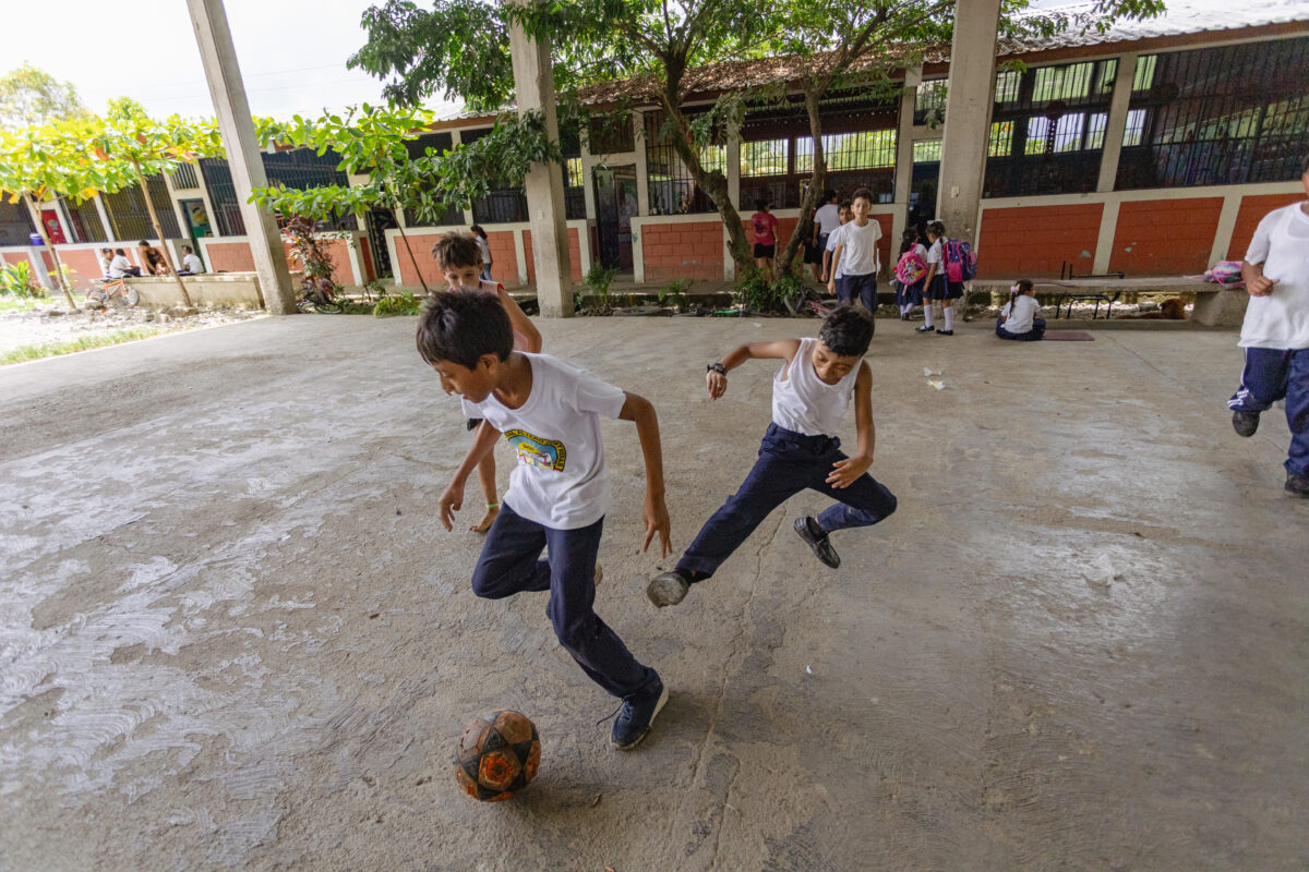 schoolchildren playing socces on a concrete courtyard. One kicks the ball while the others stand and sit nearby in the school concrete backyard