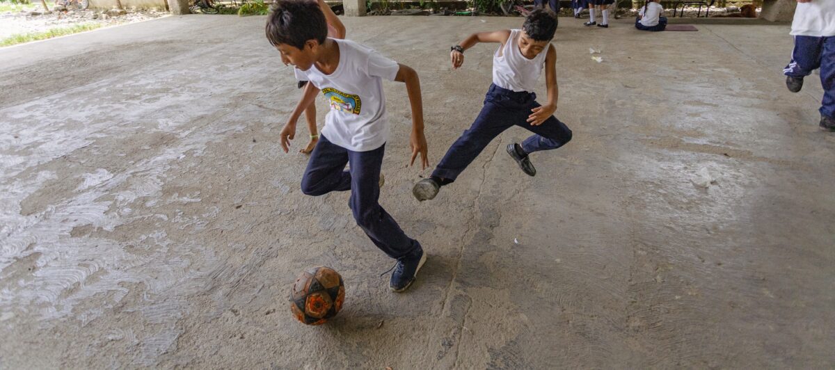 school children playing soccer on a concrete courtyard. One kicks the ball while the others stand and sit nearby in the school concrete backyard