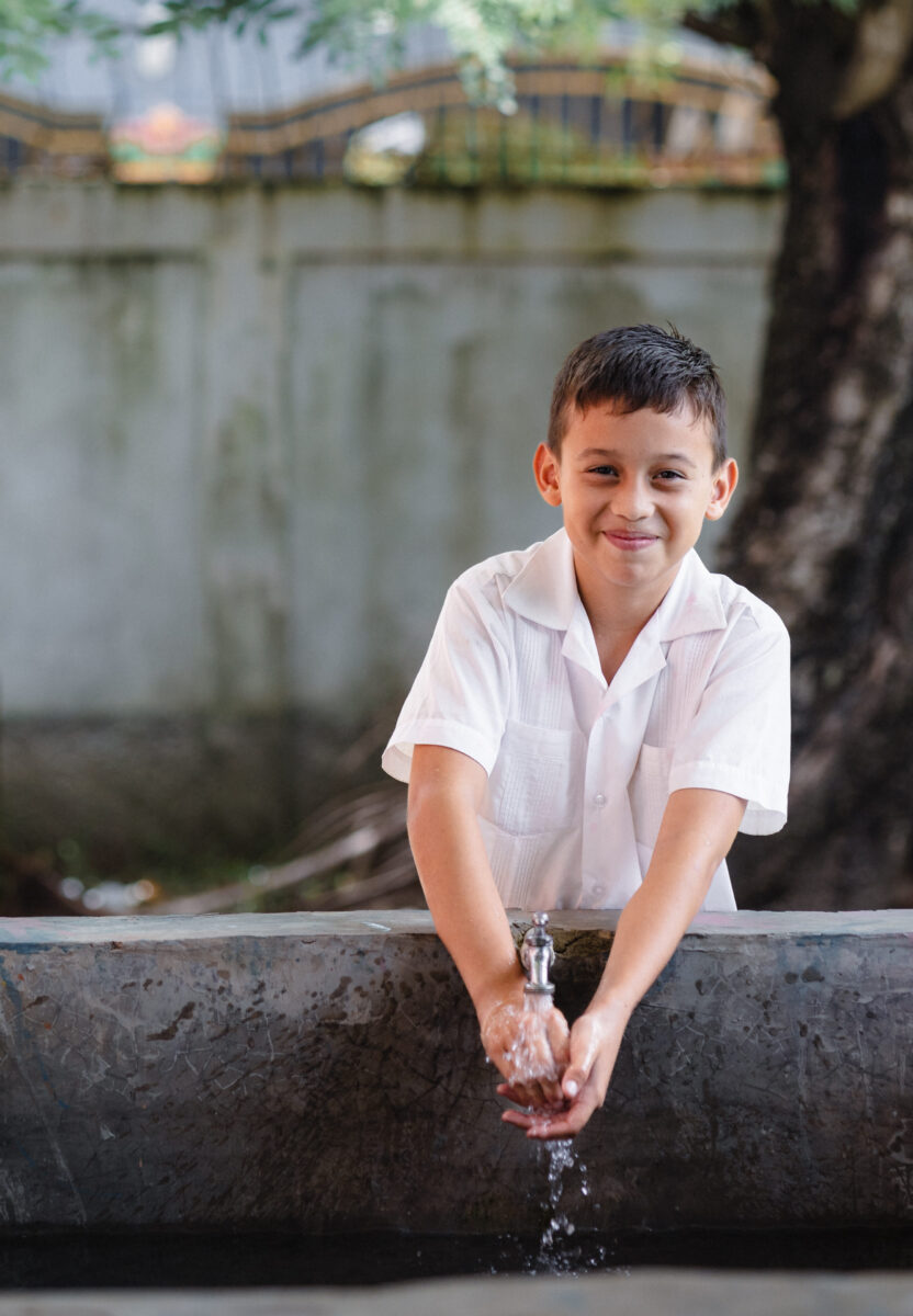 Luis washes his hands in a concrete water basin while he smiles toward the camera