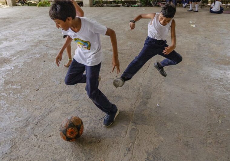 school children playing soccer on a concrete courtyard. One kicks the ball while the others stand and sit nearby in the school concrete backyard