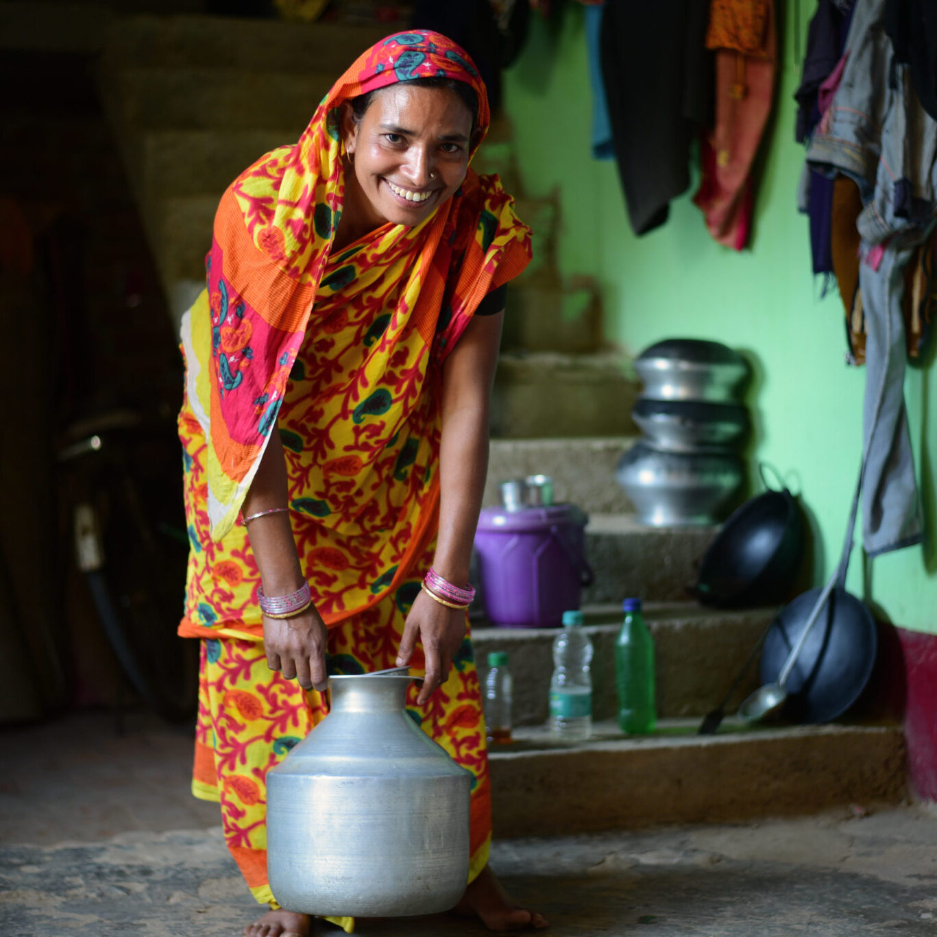 A woman in a brightly colored sari holds a container of water in her home
