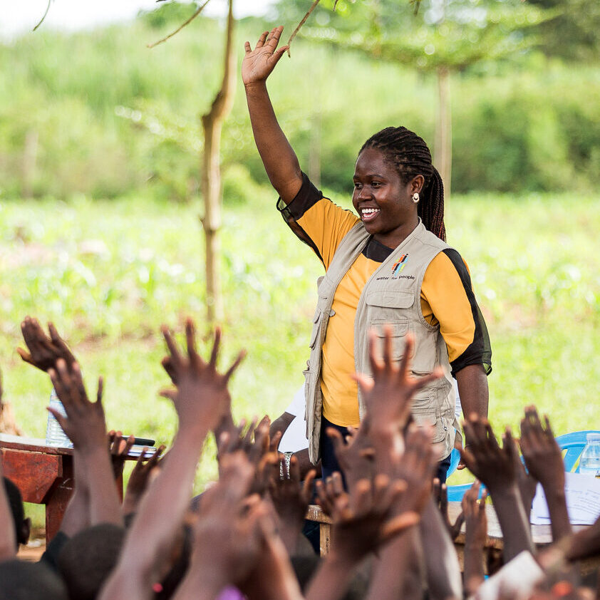 A woman wearing a Water For People vest raises her hand in front of a group of students also raising their hands against a green, outdoor background.