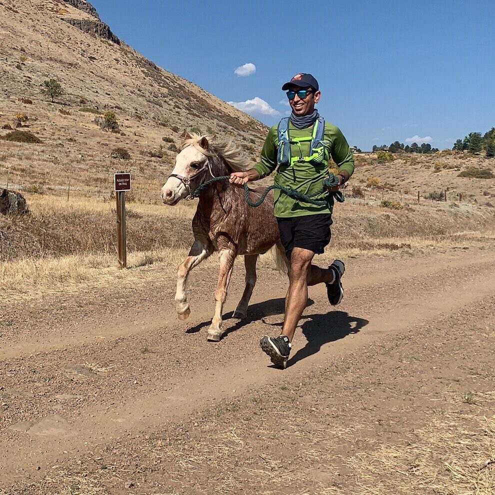 A man in green runs with a pony besides him across brown, dusty ground.