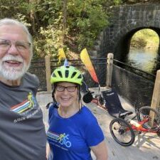 A man, left, and a woman in a bike helmet, right, in front of a bicycle and a stone bridge